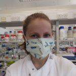 Selfie of me in a lab wearing a lab coat and mask. There's shelves of chemicals in the background.