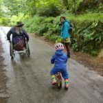 3 people walking in the woods. One is in a wheelchair, another is a toddler on a small bike, the third on foot.