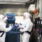 Lizzie and Rob wearing white space suits, while placing cells into a blue shredder. The shredding takes place in a safety chamber.