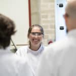 Photograph of Lizzie smiling, while wearing her lab coat and lab specs. In the foreground is the backs of her two colleagues: Rob and Anton.
