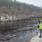 A person in a yellow safety jacket stands next to a river. They hold a long stick with a small bucket at the end to take water samples. It snows.