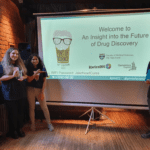 On the left Victoria and Tejal displaying Pint of Science Prizes; a pint glass and a stickman digital timer and on the right John is holding a Newcastle university chemistry department branded plastic water bladder.