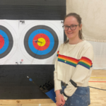 A picture of Isabelle standing next to an archery target wearing a rainbow striped jumper. She is smiling because she is very proud. She has managed to land three arrows in the center bulls eye of the target!