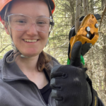 A picture of Isabelle holding her hands up in a thumbs up gesture. She is wearing safety gear such as a helmet and safety glasses. You can see the climbing rig in the photo.