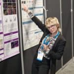 A woman with short blond hair wearing smart clothing pointing at a large poster with research data.