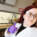 A purple-haired person wearing glasses, lab goggles, a lab coat and purple gloves gives a thumbs up in front of a chemical fume hood.