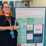 An orange haired-person, wearing glasses, a headband and a lanyard stands in front of a poster entitled "Antimicrobial Peptide Pores and Where to Find Them".