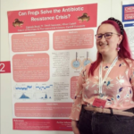 A purple-haired person with glasses stands in front of a poster entitled "Can Frogs Solve the Antibiotic Resistance Crisis?"
