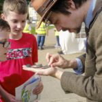 Two children looking intently at two small fossils held in the hand of a man with a hat. The man is also looking at the fossils.