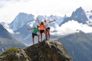 Three people stand on a summit. There are mountains in the background. 