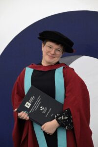 Hannah holding a printed copy of her thesis and wearing the University of Nottingham PhD graduation gown (burgundy red, pale blue trim, with a very silly floppy black hat).