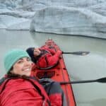 A selfie of two women dressed in red drysuits in a red kayak. next to the front of a glacier.