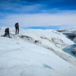 Three people walk over the surface of the ice. A bright blue river of meltwater cuts across the surface.