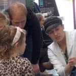 A person in a white lab coat and a fabric cap gestures to a piece of pH paper. They are describing the colour of the paper and what that means in terms of pH to 2 young girls between the ages of 5 and 8.