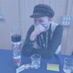 A person in a fabric hat and marine life themed waistcoat stares intently at a can of Diet Coke. There are glass beakers on the table beside them, as well as pH paper.