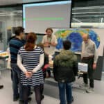 A photo of myself talking about earthquake hazards to members of the public standing in front of a world map and a live seismometer feed.