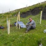 A photo of me sitting inside a fence with a solar panel and GPS unit - a completed seismometer station deployment.