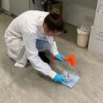 A female scientist wearing a labcoat is crouched on the floor with an orange hammer in one hand and a metal container in the other