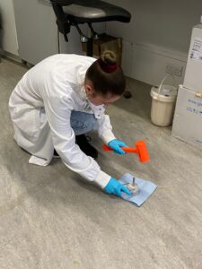 A female scientist wearing a labcoat is crouched on the floor with an orange hammer in one hand and a metal container in the other