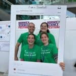 Three ladies and a man in matching green teeshirts are smiling through a selfie frame. Their teeshirts say "Ask me about cancer research"