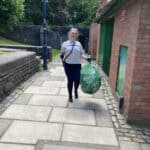 A white woman walking along a path holding a full bag of litter and a litter picker