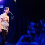 A 7 woman who is 7 months pregnant and wearing a t-shirt is standing and smiling in front of a microphone. She is presenting to an audience in front of her.