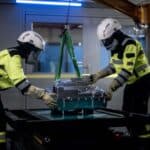 Two people moving an aluminium battery pack, using a crane. Both are wearing high-vis protective clothing and helmets.