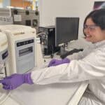 Woman in a lab coat and goggles placing a tube into a piece of lab equipment