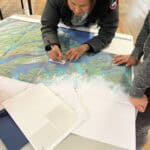 This is an image of a woman drawing on a map laid out on a table.