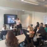 A teacher at the front of the class pointing at a screen. A class of students is watching her.