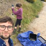 A woman with a watering can watering a bank while a man takes a selfie